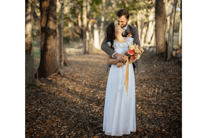 Groom holding the bride from behind while she looks up at him while they are surrounded by trees