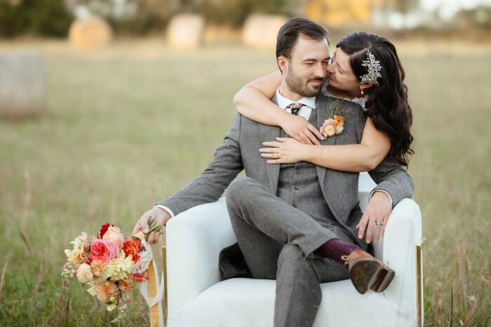 Bride hugging the groom from behind while he is sitting in a chair outside